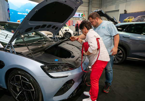 Two individuals are inspecting a light blue electric vehicle with its hood open, surrounded by other modern cars at an auto show focusing on new energy vehicles and plug-in hybrid models.