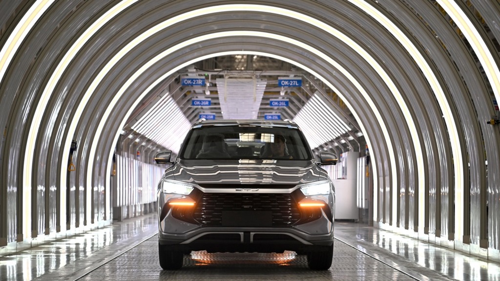A sleek, modern electric SUV is positioned inside a brightly lit, high-tech manufacturing tunnel, likely part of an assembly line for new energy vehicles such as BYD, Tesla, Geely, or Leap Motor.